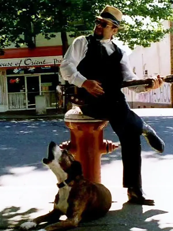 A man in a wide-brimmed hat and sunglesses sits on a fire hydrant in downtown Bellingham, playing the guitar. At his feet, a dog lies on the ground, earnestly singing along.