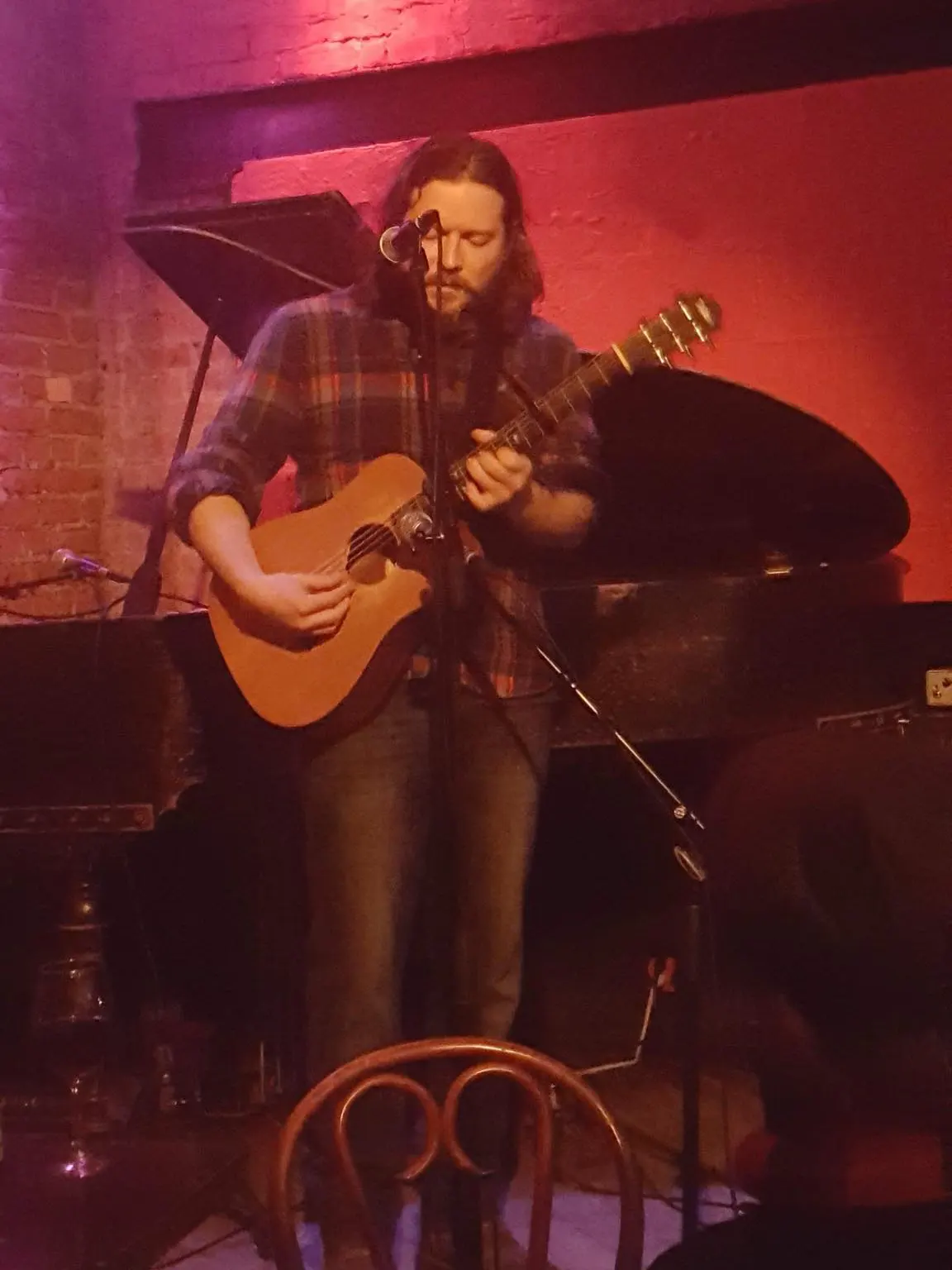 A white man with long, brown hair in a plaid flannel shirt looks down to his guitar, which he plays at a show in a venue full of multicolored lights.