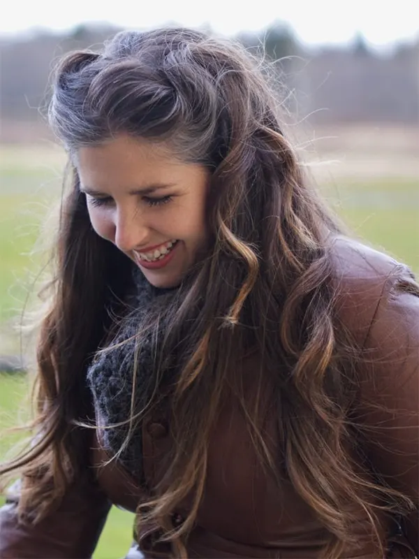 A 30ish woman with long, brown hair grins as if mid-laugh, looking down and away from the camera. She wears a brown jacket matching her hair and a black scarf, standing in front of a field distantly backed by a forest.