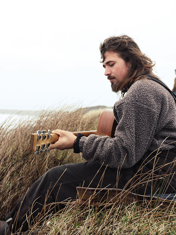 A young, windswept white man in a thick gray sweater and black pants sits on a hillside by the sea, pensively playing a guitar.